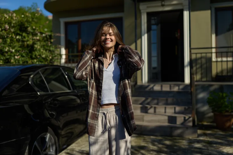 Bright morning scene shows a smiling female in a plaid jacket and white top standing by a car in front of a modern house, conveying a relaxed, confident lifestyle
