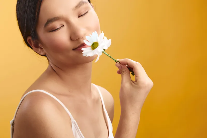 Asian woman in a light outfit smiles softly as she inhales the scent of a white daisy, set against a warm yellow backdrop, conveying calm wellness and gentle joy.