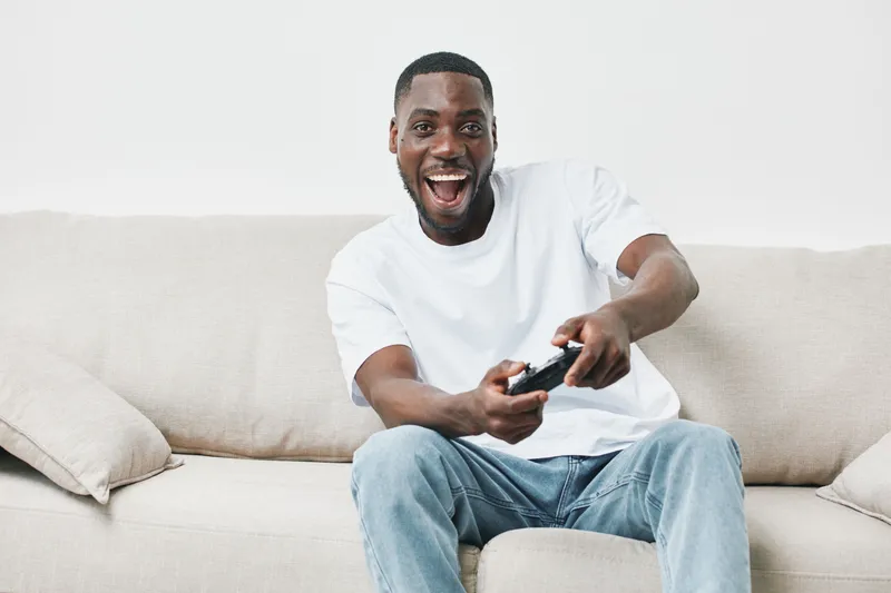 Happy young man playing video games on the couch in a bright, modern living room, wearing a white t shirt and jeans, expressing excitement and joy