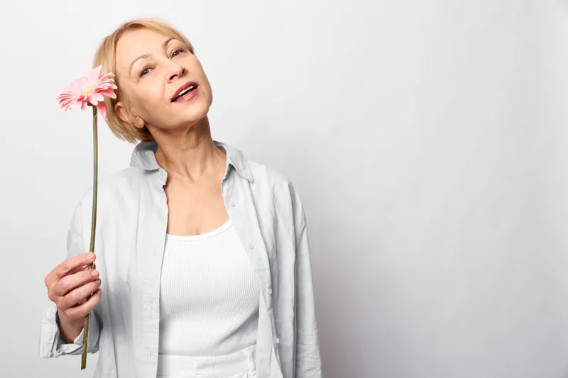 Woman holding feather on white studio background, allergy and sneezing concept, respiratory health for senior adults, closed eyes show discomfort and gentle relief