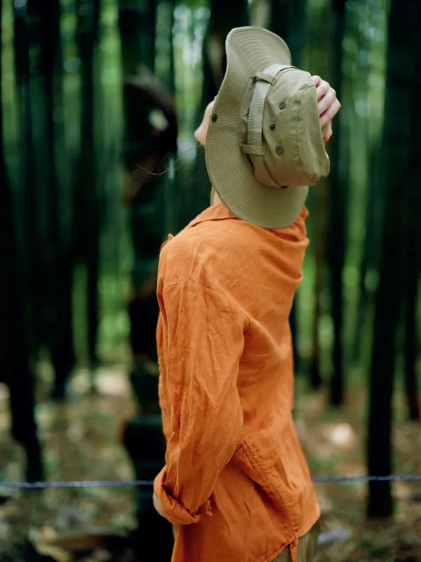 Person in orange shirt with hat seen from back in a dense bamboo forest, a nature travel portrait of a traveler standing among green trees and sunlight, serene woodland exploration mood.