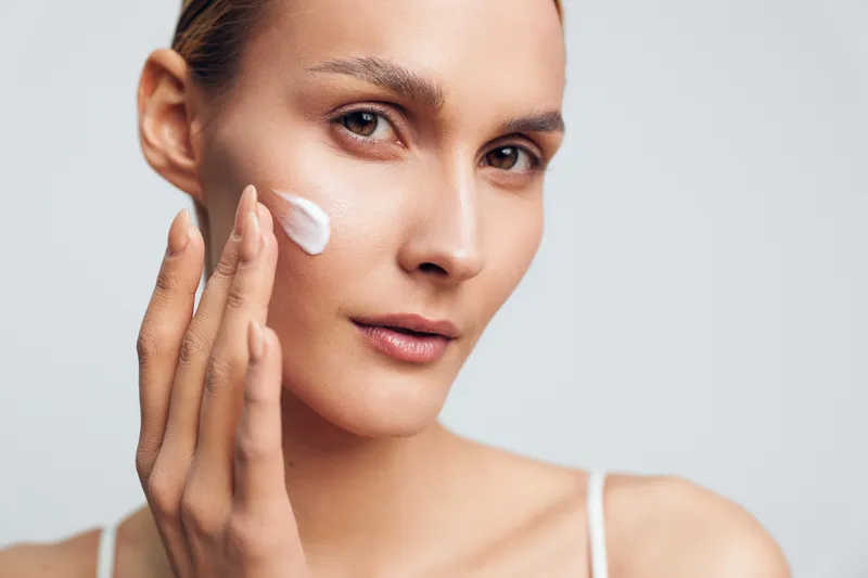Woman applying white facial cream on her cheek. Close up portrait with soft lighting and a neutral background. Skincare, wellness, and self care themes emphasized in a calm, clean mood.