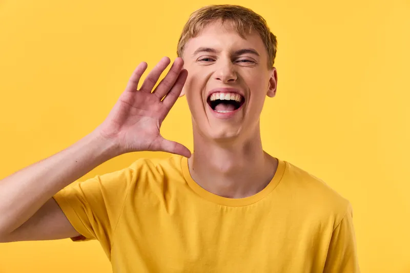 Shouting, energetic man in a bright yellow shirt greets with a wide smile, hand near his mouth, creating a sense of enthusiastic communication, promotion, and lively vibe ideal for upbeat marketing