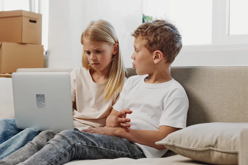 Two children sitting on a couch using a laptop together, focused and engaged in learning or entertainment at home in a bright living room setting.