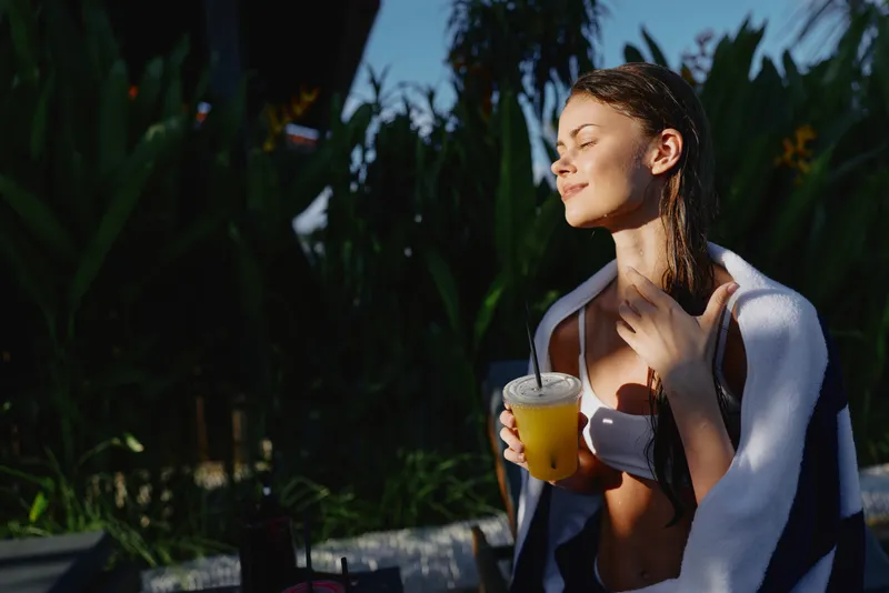 Relaxed woman outdoor enjoying a beverage under warm sunlight, towel draped over shoulders, bikini top, conveying wellness, self care and leisure in a sunny scene. Bright atmosphere.