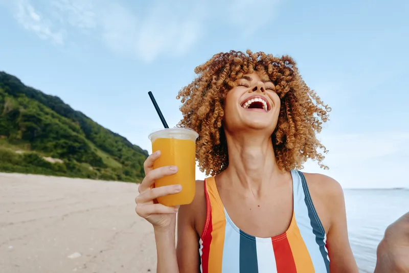 A young woman with curly hair laughs joyfully while holding a cup of orange juice on a sunny beach with green hills in the background.