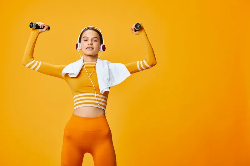 Woman fitness athlete lifts dumbbells with arms raised, wearing orange outfit and a white towel over shoulders, against a bright orange background, showcasing a dynamic workout vibe in a studio