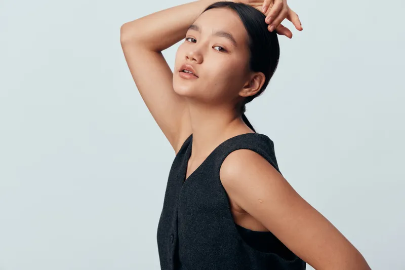 Woman posing with hand on head and wearing sleeveless black top on plain white background. Asian model with natural makeup and sleek hairstyle in studio portrait.