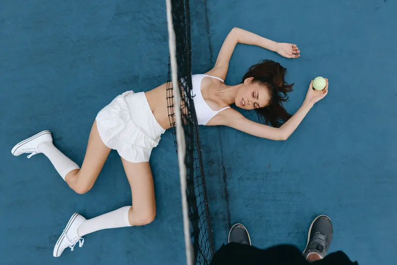 Young female athlete exhausted on tennis court, wearing sports attire, expressing fatigue, surrounded by blue tones, concept of sports dedication and effort