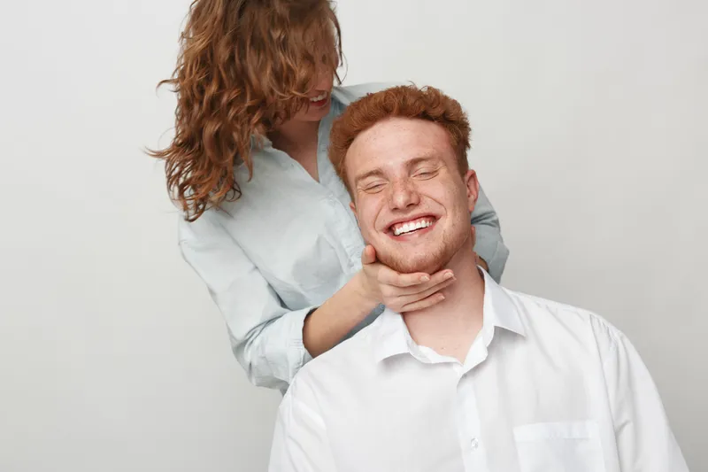 Happy young man with red hair smiles with closed eyes while woman with curly hair gently holds his face. Concept of affection, friendship, joy, and positive emotions in casual setting.