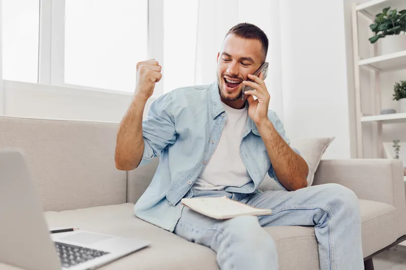 Happy man celebrating success on a phone call, wearing casual light blue clothing, seated comfortably in a modern living room with a bright atmosphere.