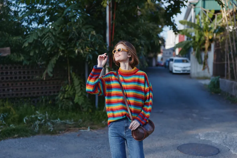 Woman in her twenties wearing a rainbow sweater and sunglasses, walking and smiling on an urban street in a casual outfit with crossbody bag, relaxed travel lifestyle portrait.