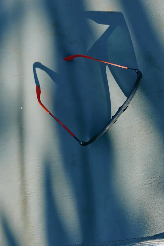Sunglasses hang on a textured wall with soft shadows, creating a casual lifestyle moment. Minimalist composition, warm light, and relaxed mood evoke summer days and everyday style.