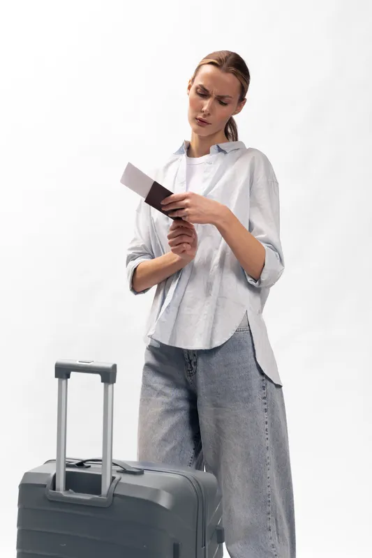 Young woman with suitcase holds passport and ticket looking worried or confused. Travel concept with casual clothing and white background. Preparing for trip or checking documents.