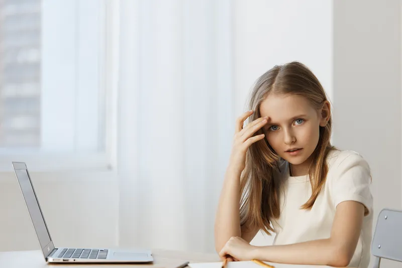 Pensive young girl with long hair in a white dress sitting at a desk with a laptop, depicting stress and concentration in a bright office environment