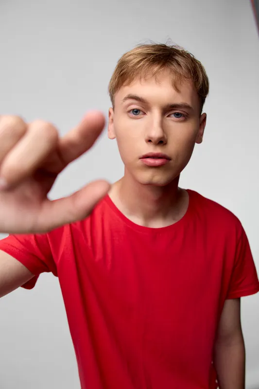 Close up of a young man in a red shirt reaching toward the camera, hand in foreground, focused gaze and intense expression, simple studio backdrop, dynamic composition conveying immediacy and