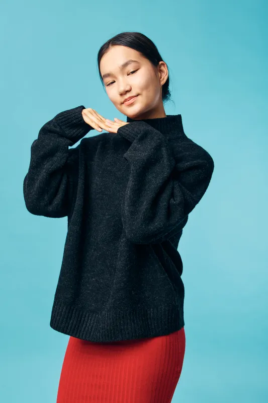 Young woman in black sweater and red skirt posing with closed eyes and gentle smile on blue background. Relaxed female model showing calm and peaceful expression with hands near face.