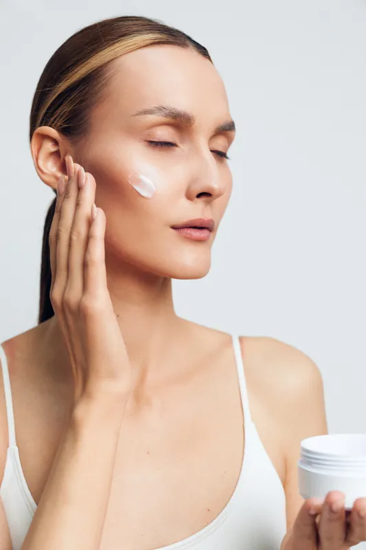 Young woman with light skin applies facial cream to her cheek in a clean studio, calm expression, soft lighting, natural tones, minimal background, white tank top