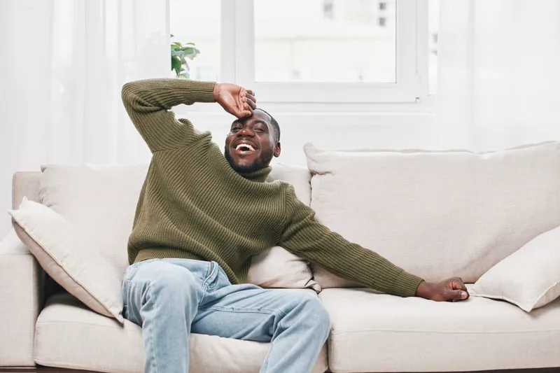 Happy young Black man in green sweater laughing on cozy sofa, enjoying his day at home with bright natural light and relaxed atmosphere