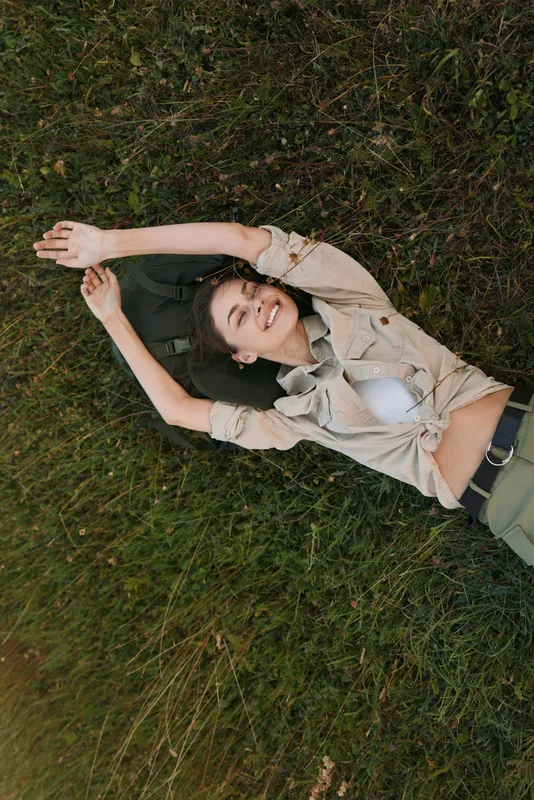 Young woman lies on grass with arms overhead, wearing a beige shirt and light top, enjoying a relaxed outdoor moment.