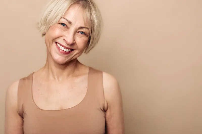 Smiling middle aged woman with short blonde hair wearing beige top poses against warm beige background. Concept of natural beauty, aging gracefully, confidence and positive emotions.
