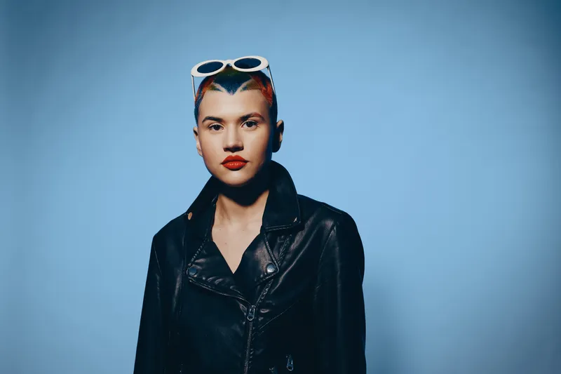 young woman with short colorful hair and sunglasses wearing black leather jacket standing against blue background with confident facial expression