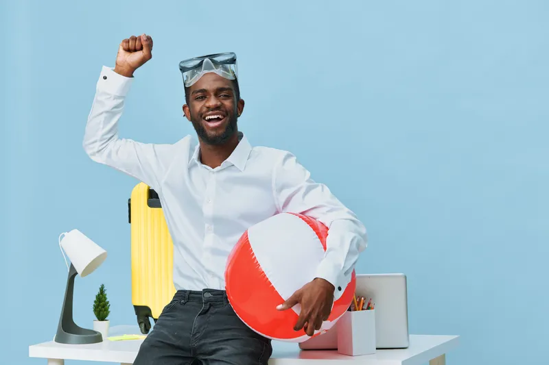 Happy young man with dark skin and beard in casual white shirt holding a red and white beach ball, celebrating at workspace with travel gear and office supplies, cheerful mood, summer vacation concept