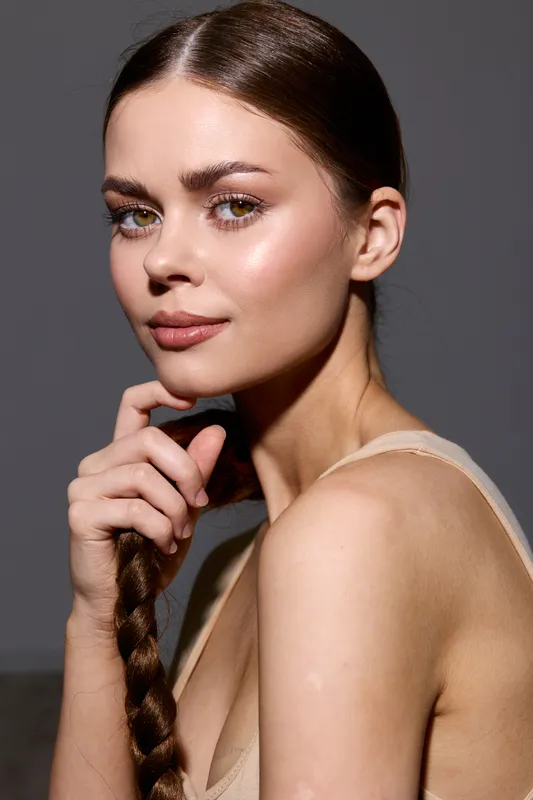 beauty woman with braided hair and glowing skin poses confidently against a neutral background in soft natural light, showcasing subtle makeup and smooth complexion.