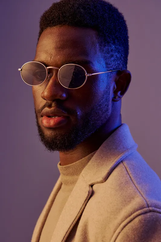 Stylish young Black man wearing round glasses and a beige coat, showcasing a confident expression against a soft purple background