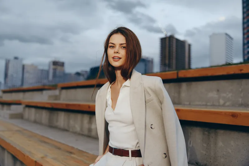confident woman in beige coat and white blouse sitting outdoors in urban environment with modern buildings background autumn fashion business style