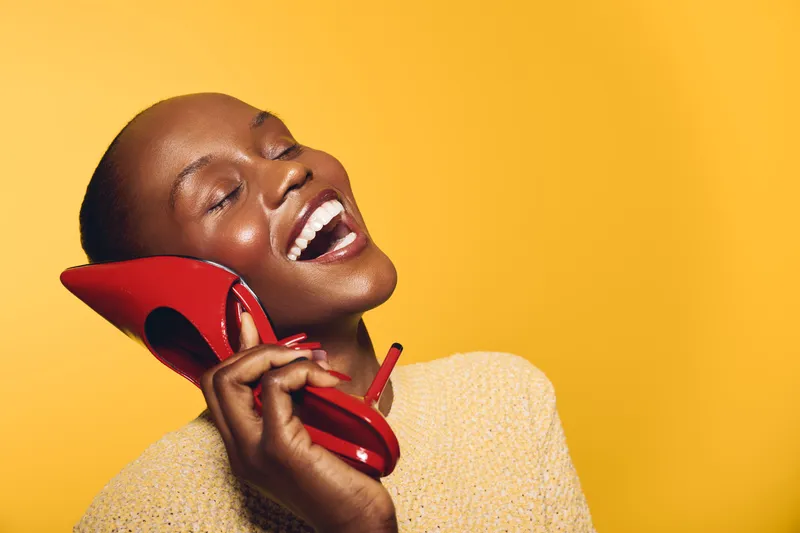 Happy African American woman with dark skin laughing and holding red high heel shoe near ear as phone on yellow background. Concept of joy, fashion, and playful communication.