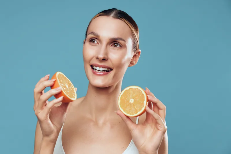 Young woman with brunette hair holds two halves of an orange near her face while smiling against a blue background, conveying freshness and healthy lifestyle.