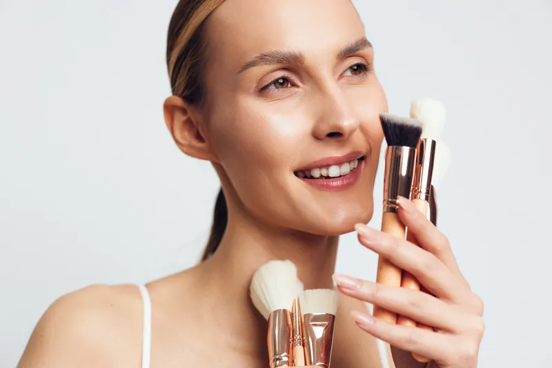 A smiling woman holds makeup brushes near her face, showcasing cosmetics and skincare vibes. Close-up portrait focused on beauty routine and a fresh look