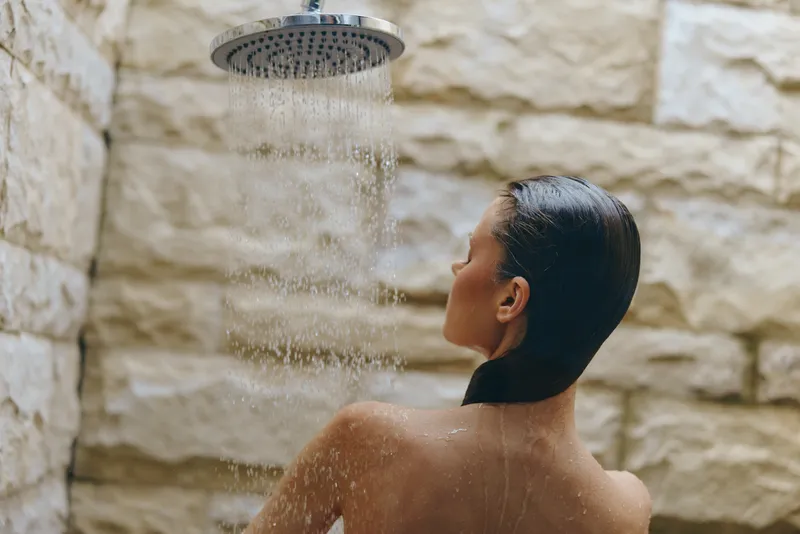 Woman enjoying outdoor shower with water falling, natural stone background, wet hair, relaxation, wellness, summer vibe