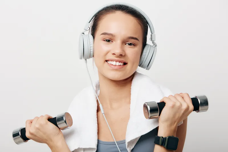 A young woman in fitness gear lifts dumbbells with headphones on and a towel draped around her neck, smiling after a workout in a bright gym setting.