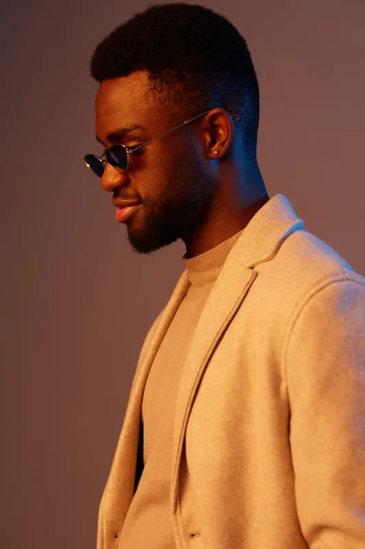 Stylish young man in trendy sunglasses, wearing a neutral colored blazer, exuding confidence against a soft blue and beige background, ideal for fashion and lifestyle concepts