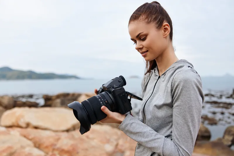 Young woman in sportswear examining camera at the coastline, enjoying nature and photography, reflecting vibrant outdoor lifestyle