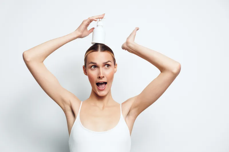 Young woman in a white tank top balances a white bottle on her head with a playful and surprised expression against a plain white background.
