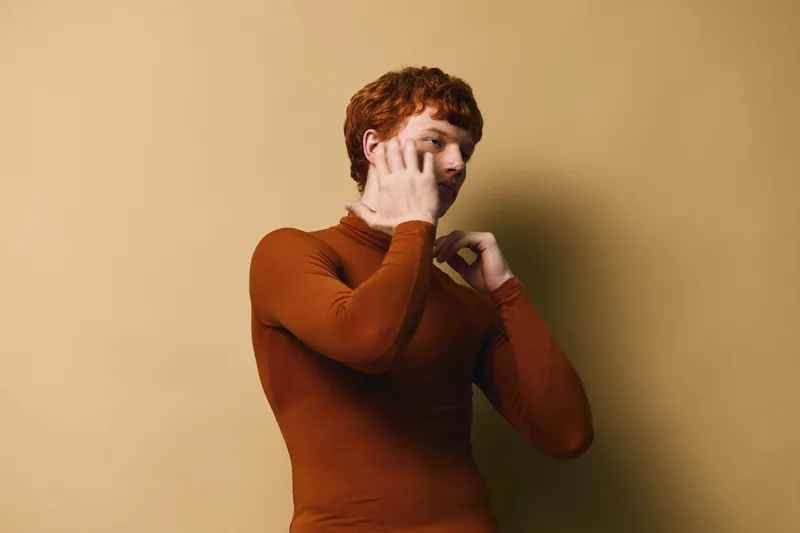 Young man with red hair in brown sweater talking on smartphone against beige background, casual communication and technology connection concept with thoughtful expression.