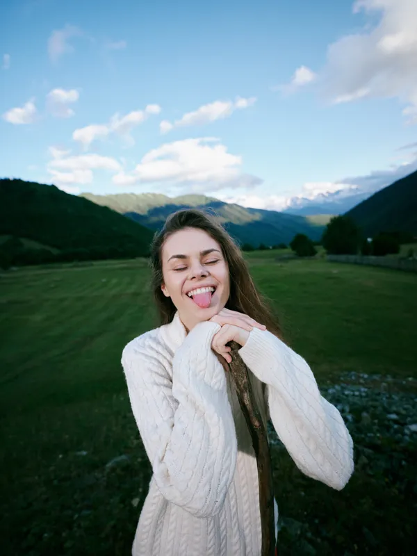 Playful woman outdoors, sticking tongue out in a cozy white sweater, surrounded by green hills and blue sky, capturing carefree mood, fresh air, and natural charm during a joyful mountain retreat