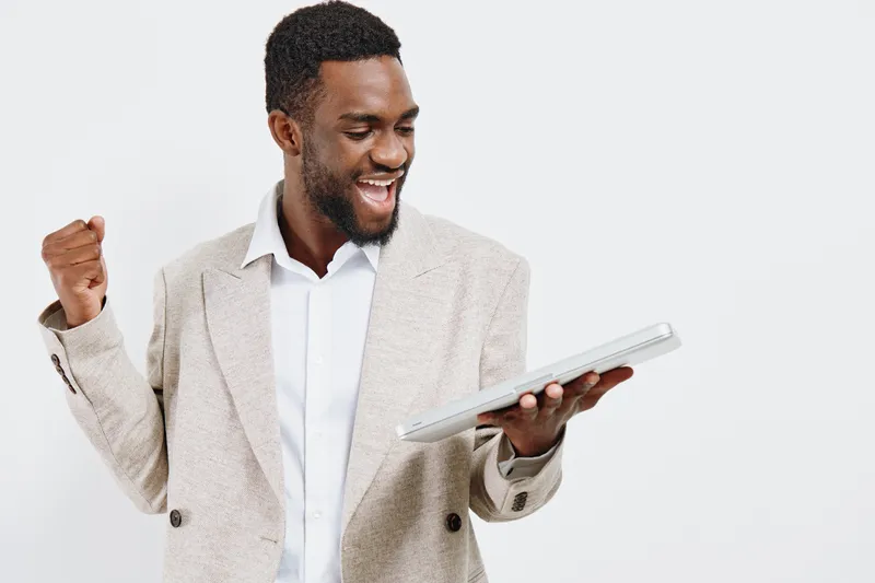 Smiling happy young African American man in beige blazer holding tablet device isolated on plain white background studio portrait. People lifestyle concept