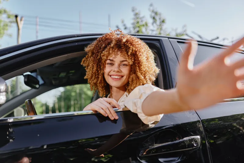 smiling woman with curly hair sitting in car window, bright natural light, casual white blouse, sunny day, cheerful mood, urban background