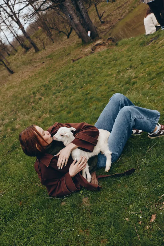 Woman lying on grass in a park, hugging a white goat kid, casual clothes. Intimate moment of affection, outdoors, soft natural light, rustic mood. Story of companionship, playfulness, and simple