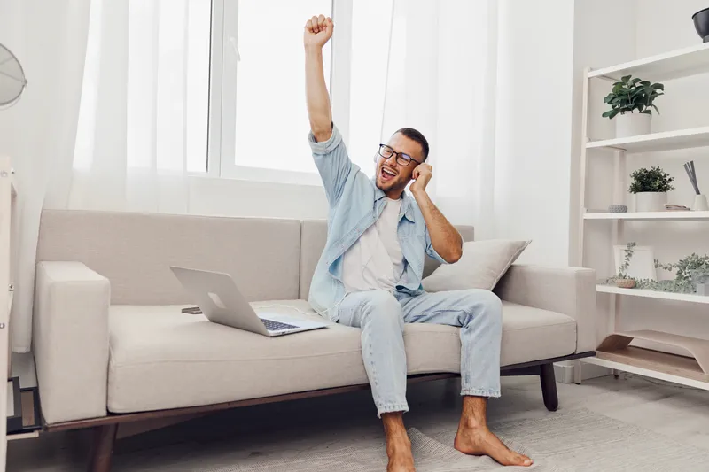 Happy man celebrating a work achievement at home, embracing digital detox and self-care with emotional openness and vulnerability in a bright, cozy setting.