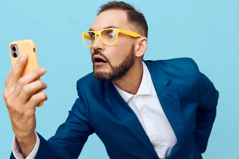 A focused business professional man, wearing a blue suit and yellow glasses, is captured against an isolated blue background, conveying surprise and curiosity while checking a smartphone.