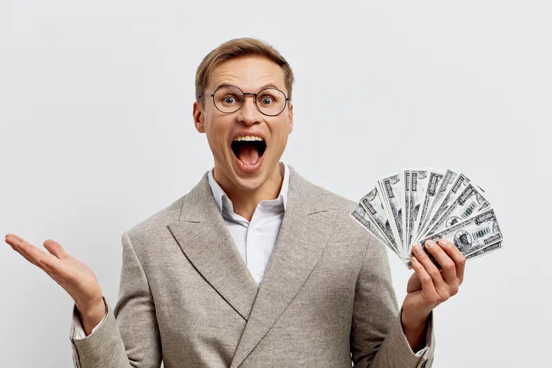 Excited happy man with glasses, wearing a beige suit, holding a fan of dollar bills, expressing surprise and excitement, isolated on a plain white background, studio portrait. People celebration conce