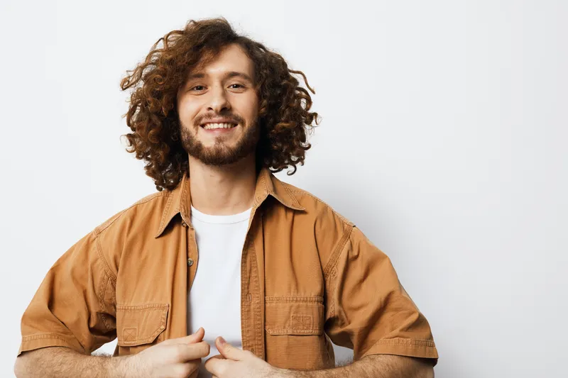Smiling young man with curly hair in a casual outfit, standing against a light background, showcasing a joyful emotion and vibrant style
