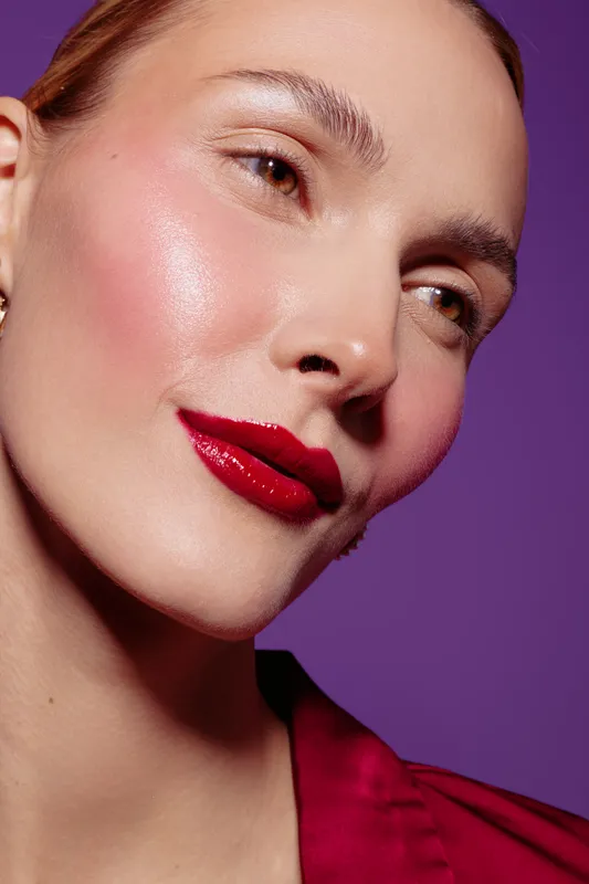 Close-up of a woman with glossy red lips and glowing skin on purple background. Makeup and beauty concept with natural light highlighting facial features and smooth complexion.