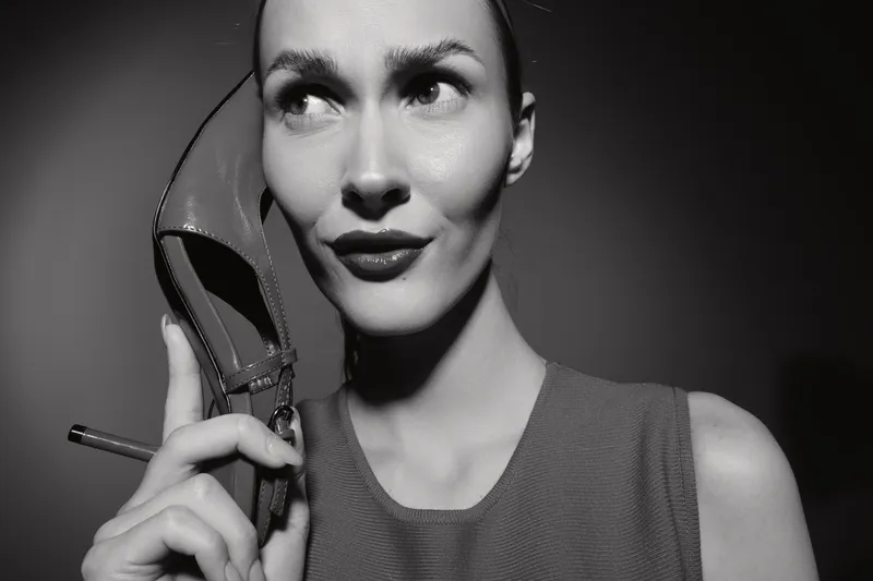 Portrait of a light skinned caucasian woman holding a high heel shoe and a cigarette near her face. Black and white studio lighting creates a dramatic vintage mood.