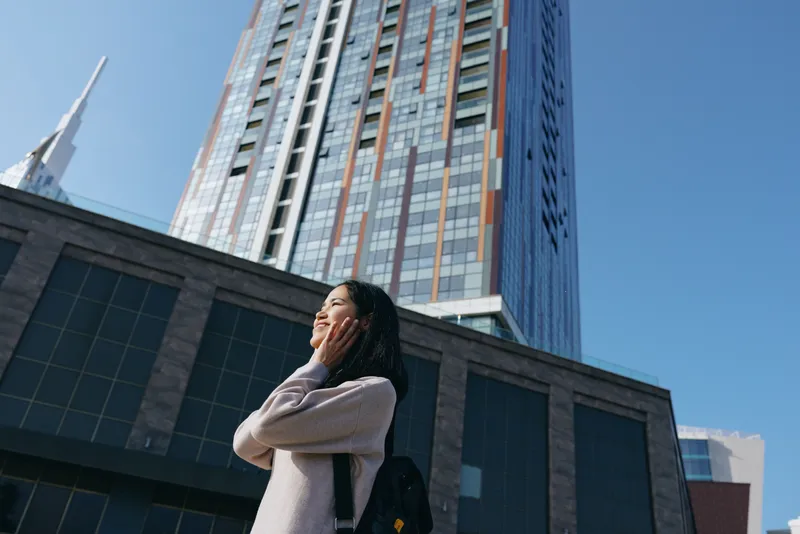 Woman with backpack gazes up at a towering skyscraper in a sunlit urban plaza, capturing ambition and curiosity amid modern city architecture and travel